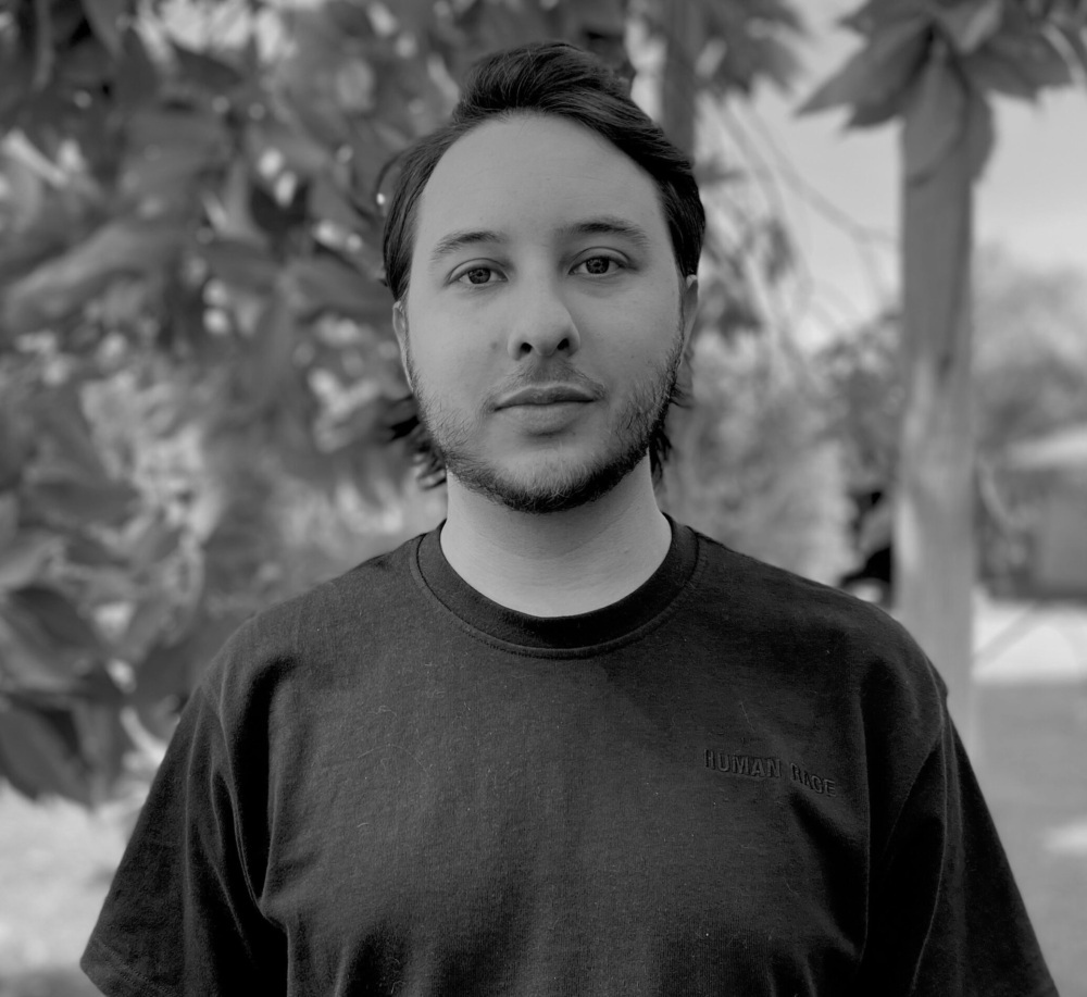 Black and white photograph of a man standing in front of shrubbery, looking into the camera, smiling, and wearing a dark-colored shirt.