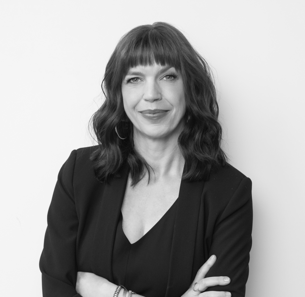 Black and white photograph of a woman looking into the camera, smiling, wearing a dark-colored jacket over a dark-colored shirt, and with her arms crossed.