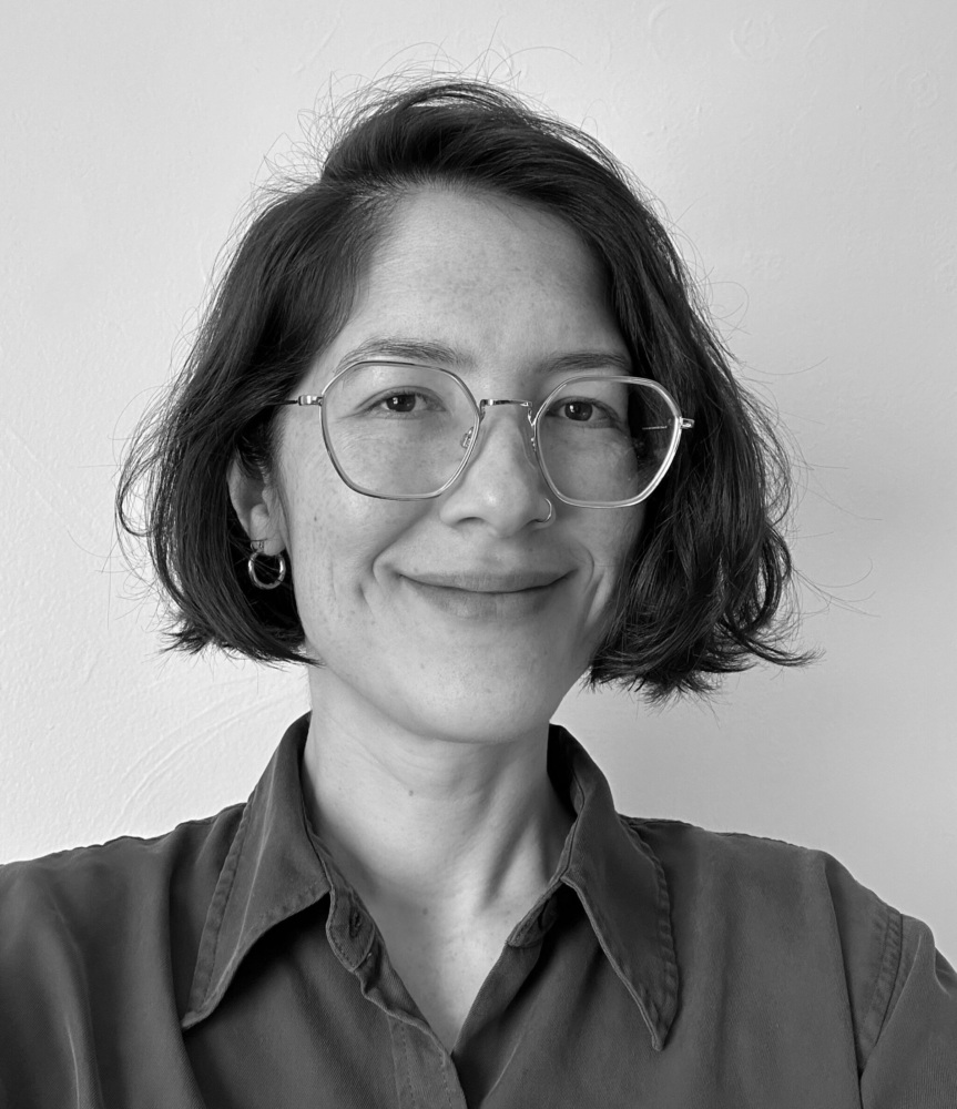 Black and white photograph of a woman looking into the camera, smiling, and wearing glasses and a dark-colored collared shirt.