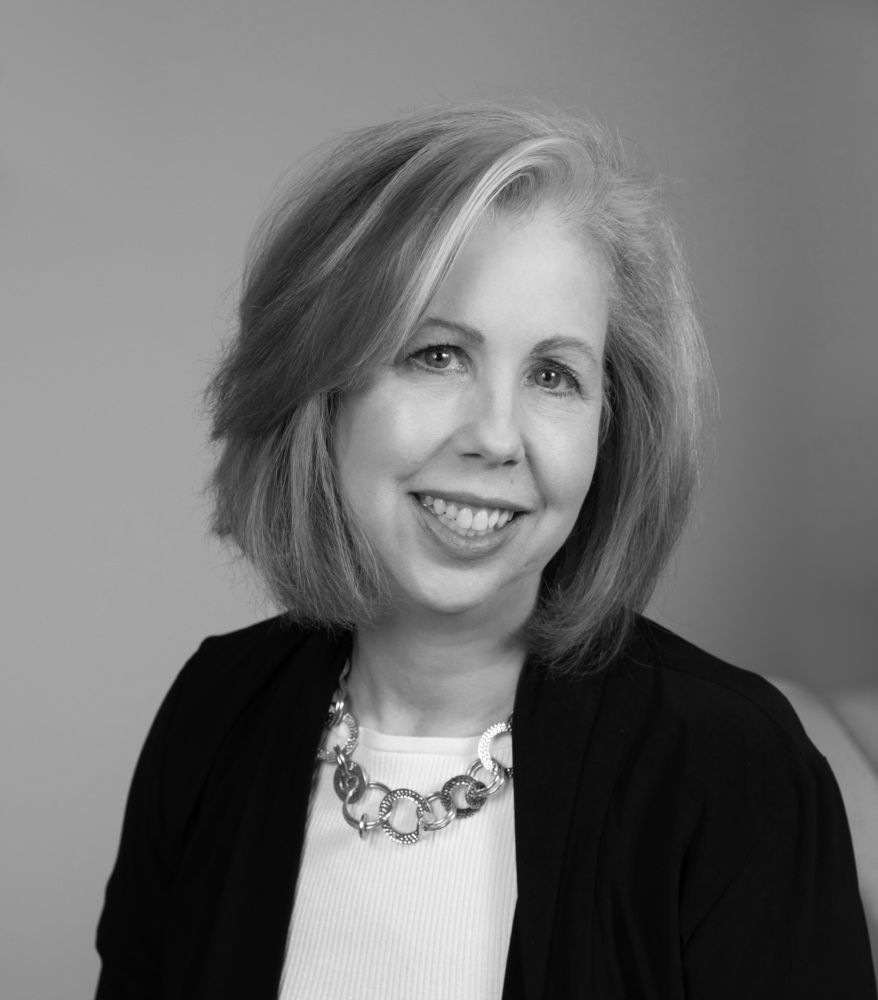 Black and white photograph of a woman looking into the camera, smiling, and wearing a dark-colored jacket over a light-colored shirt and necklace.